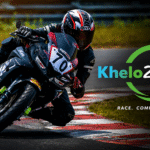 What is a Racer Bike leaning aggressively through a racetrack corner with the Khelo24Race logo, showcasing high-speed motorcycle racing and professional track performance.